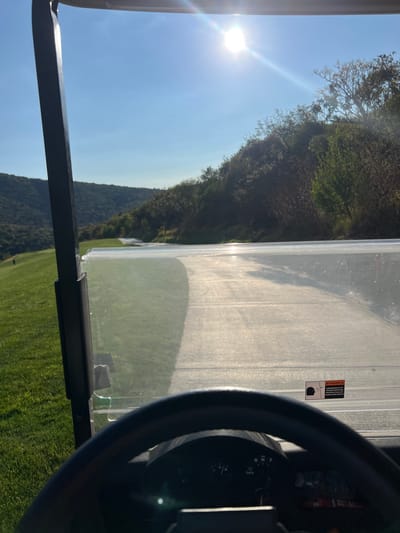 The image depicts a golf course view from inside a golf cart. The sun shines brightly above green trees and grassy hills visible through the windshield. The cart's frame, steering wheel, and dashboard controls are partially visible in the foreground. A safety warning sticker is affixed to the windshield.