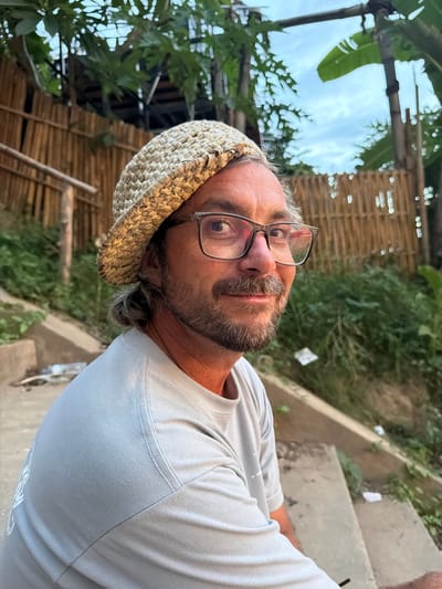 A man wearing a straw hat and glasses is pictured outdoors. He has a beard and his hair is tied back in a bun. He is wearing a light-colored t-shirt. The background shows a bamboo fence, lush green foliage, and a cloudy sky. There are concrete steps in the foreground. The image appears to have been taken in Luang Prabang, Laos, given the architectural style of the fence and the tropical vegetation.