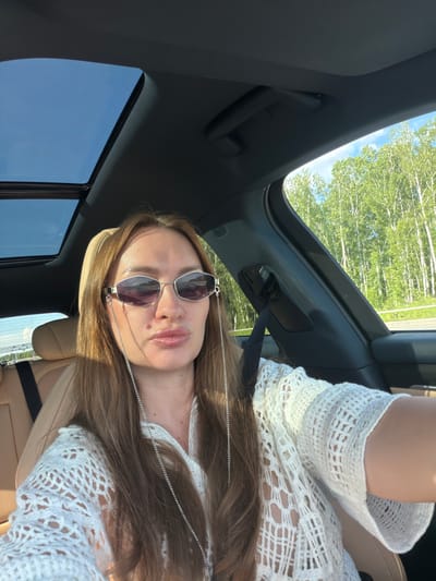 A woman with long brown hair and sunglasses is sitting in the passenger seat of a car, taking a selfie. She is wearing a white crocheted top and a silver chain hangs from her sunglasses. The car has a sunroof that is open, revealing a blue sky. Outside the car, there are trees and a road. The image was taken in Severny, Russia, on a sunny day.