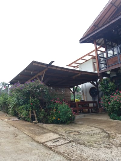 A two-story building with a wooden balcony stands near a covered patio. The patio's roof is corrugated metal, supported by wooden posts, and features bamboo screening at the back. A concrete pathway leads to the structure, bordered by dense foliage and colorful flowers. The sky is overcast.