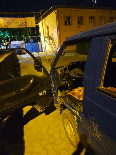 A dark blue van with its driver's side door open is parked at night on a paved surface in Panjakent, Tajikistan. The van shows signs of wear and tear, including scratches and damage to its exterior. The interior of the van is visible, revealing the steering wheel, dashboard, and a beaded seat cover. A yellow sign with text in an unfamiliar language is suspended above and to the left of the van, near a building with yellow walls and an outdoor staircase. The lighting suggests artificial illumination, possibly from streetlights or building lights, casting yellow hues and shadows. The environment appears to be a street or parking area in a semi-urban setting.