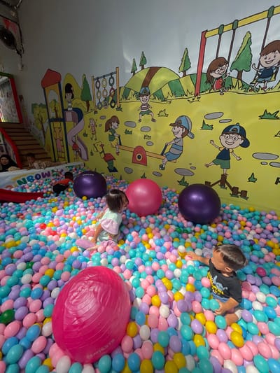 The image depicts an indoor playground in Sinduagung, Indonesia. The space features a mural of children playing in a park painted on a yellow wall. The primary feature is a ball pit filled with multi-colored plastic balls. Two children, a boy and a girl, are in the ball pit. The boy is wearing a black shirt and the girl a light-colored top. Several large exercise balls in purple and pink are also present in the pit. A red slide is visible near the edge of the pit. Several people, including women, are standing at the edge of the play area. A doorway and other parts of the play structure are visible in the background.