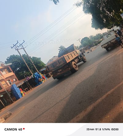 The image captures a daytime street scene in Obe, Nigeria, under a bright, clear sky. The asphalt road, slanting from the lower-left to the upper-right, is the primary subject. A dark, rusted dump truck, displaying the number "50" on its rear, occupies the midground, oriented away from the viewer. Further along the road, a white pickup truck is visible, also moving away. An individual is positioned near the white pickup truck on the right side of the road. On the left, a two-story building with a reddish-brown facade features clothes hanging from a balcony. Adjacent to this building are smaller structures with a retail setup, characterized by numerous stacks of colorful plastic crates (blue, red, yellow, green). Multiple wooden utility poles with overhead power lines parallel the road on the left. Vegetation, including trees, is present in the background and along the roadside. Long shadows are cast on the road surface from the left.