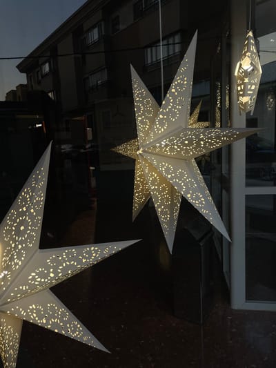 This is a close-up, angled outdoor shot of a storefront window display featuring decorative, illuminated paper stars. The stars are white with intricate cut-out patterns that allow light to shine through, creating a warm glow. In the background, a dark, multi-story building with windows is visible, reflecting the twilight sky. Some reflections of cars and interior lights are also present in the window. The scene is set in La Línea De La Concepción, Spain, a city on the easternmost edge of the Campo de Gibraltar, bordering Gibraltar.