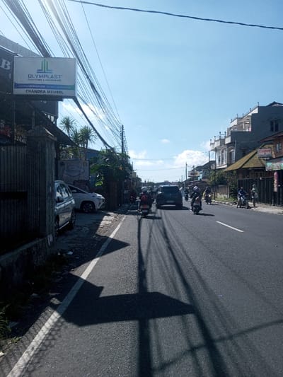 The image depicts an urban street scene in the city of Ban Phonhxavanh, Laos, under a clear blue sky with bright sunlight, casting long, dark shadows across the asphalt road. On the left side, a gray metal fence runs along a sidewalk and parking area, where a silver sedan and a white SUV are parked. Above, a rectangular backlit sign displays "OLYMPLAST FURNITURE & HOUSEWARE" and "CHANDRA MEUBEL." A dense network of black overhead utility lines crisscrosses the upper left portion of the sky.

The two-lane road features a solid white line marking the left edge and a dashed white line separating lanes further right. Multiple individuals are riding motorcycles or scooters, predominantly moving away from the viewer, with many wearing helmets. A dark SUV is also visible in the distance, traveling in the same direction.

On the right side of the street, a row of multi-story buildings with varying architectural styles lines the road, some featuring balconies. One building prominently displays a "Canon" sign. A few individuals are visible on the sidewalk near these buildings. The environment suggests a bustling commercial area under strong daylight conditions.