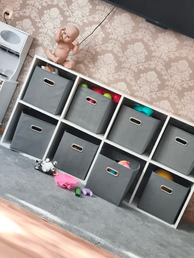 In a room with a floral wallpaper and a wooden floor, a white cube shelf unit is filled with eight gray storage bins. On top of the shelf unit, a baby doll is posed sitting with its hands on its head. Scattered on the gray carpet in front of the shelf are a stuffed husky toy, a pink doll dress, a green toy block, and a purple toy controller.