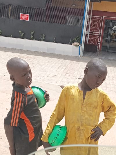 Two young males, appearing to be children, are depicted standing outdoors in Hotoro, Nigeria, during daylight hours. The subject on the left wears a black short-sleeved shirt with orange vertical stripes on the sleeves and holds an empty green plastic bowl in his left hand, looking slightly upwards and left with his mouth open. The subject on the right, slightly taller, wears a soiled yellow long-sleeved garment and holds a green plastic bowl with "M.B.C.Y. U.S.M.A." text in his left hand, looking downwards and to the right. Both individuals have short, dark hair. They stand on a surface comprising light-colored interlocking pavers mixed with bare dirt patches. In the background, to the left, is a dark grey building facade with vertical slats, a white low wall, and sparse vegetation. A red sign is affixed to this building, reading "VEHICLES PARKED AT OWNERS RISK." To the right, an adjacent building displays a red and yellow facade, featuring a doorway with a decorative metal security grate and reflective glass, showing a partial, blurred reflection.