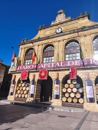 This is a photograph of the front of the Haro Town Hall in Haro, Spain, a city renowned for its wine production. The building is constructed of stone and features classical architectural elements, including arched windows and decorative stonework. Above the main entrance, a large banner reads "HARO CAPITAL DEL RIOJA" (Haro Capital of Rioja), indicating the city's importance in the Rioja wine region. The banner is flanked by two large, stylized depictions of wine bottles. Below the banner, an archway is adorned with several red banners and flags, including the Spanish flag. In front of the archway, a collection of wine barrels are stacked, some bearing labels of local wineries. The ground in front of the building is paved, and shadows from nearby structures fall across the scene, suggesting it is daytime. The sky is a clear, bright blue. There are no people visible in the image.