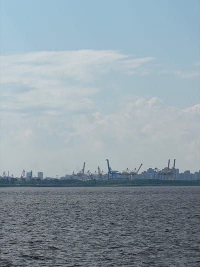 This is a wide shot of the Gulf of Finland in Saint Petersburg, Russia, on a cloudy day. In the foreground, dark, choppy water fills the bottom half of the frame. The background features a distant city skyline dominated by numerous industrial cranes and buildings, suggesting a port or industrial area. The horizon is a thin line of trees separating the water from the city. The sky is a pale blue with scattered white clouds. There are no people or animals visible in the frame.