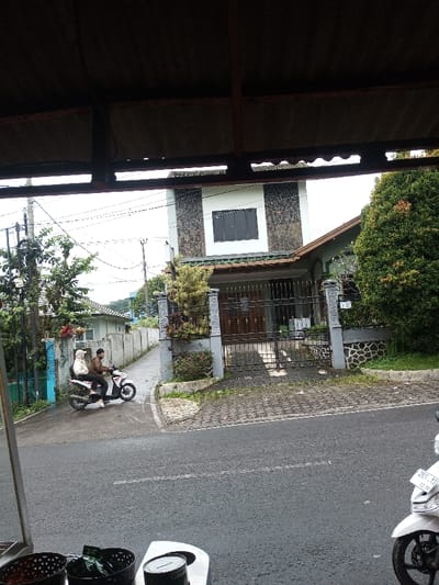 The image depicts a street scene in Cipendawa, Indonesia, viewed from beneath a dark corrugated roof structure. In the immediate foreground, partial views of two dark woven baskets, a lidded white container, and the front wheel and fairing of a white scooter (license plate D 3927 YTI 03-26) are visible. An asphalt road with faint white markings extends across the mid-ground. On the left side of the road, two individuals ride a white scooter at the mouth of a concrete alleyway; the male driver wears a brown jacket, and the female passenger a light headscarf and jacket. Across the road, a two-story residential house is prominent, featuring a lower ornate dark metal gate flanked by decorative grey pillars and a patterned stone driveway. The upper story has a light-colored facade with dark stone cladding on either side of a large dark-framed window. A green-tiled section covers the lower entrance. Dense green foliage flanks the house. The alleyway on the left extends past a concrete wall, revealing further residential buildings and utility poles with numerous power lines against an overcast sky.