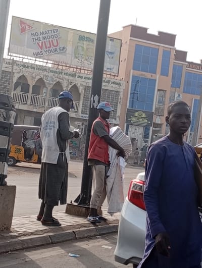 Two men are standing on a sidewalk, one wearing a vest and holding papers. Another man in a red vest holds folded white items. A man in a blue robe walks past a white car. A yellow vehicle is parked near them. Above, a billboard advertises "VIJU" drinks and another possibly "BBSTAR." A building with "AISIA FOUNDATION SCHOOL" is visible, and another labeled "TANKULL." A utility pole is near the men on the sidewalk.