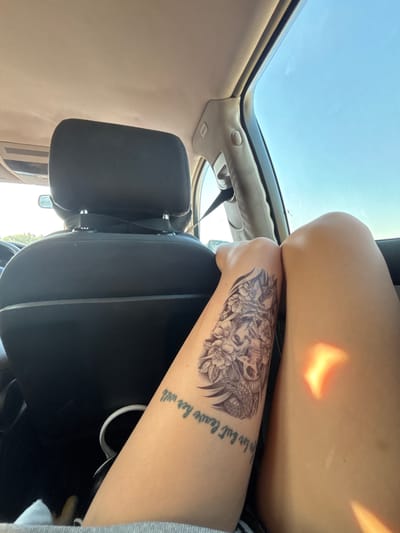 The image shows the interior of a car, looking towards the back seats and out the window. The focus is on a person's arm and legs, adorned with a tattoo of a skull entwined with flowers and a snake. Below the tattoo, there is text written on the skin, which appears to be a quote. The person is sitting in the car with sunlight casting shadows on their legs. The background shows the car's headrest, the car's interior trim, and a bright blue sky with some trees visible through the window. The image is taken from a perspective inside the car, looking backwards and slightly to the side. The location is stated as Strumyani, Bulgaria.