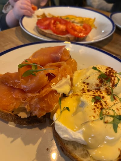 This image depicts an indoor dining scene, likely within a cafe or restaurant located in Oxford, United Kingdom. In the foreground, two breakfast or brunch dishes are prominently displayed on white ceramic plates with distinct blue rims, resting on a light-colored wooden table. The closest plate features two preparations on toasted bread: the left side contains generously arranged folds of orange-pink smoked salmon, garnished with small dollops of a white or pale-yellow sauce and a few green microgreens. The right side holds two poached eggs, thoroughly covered in a glossy, bright yellow hollandaise sauce, which is sprinkled with reddish-brown powder (consistent with paprika or chili) and further garnished with green microgreens. In the blurred background, a similar plate is visible, holding toast topped with sliced red tomatoes and a portion of yellow food, appearing to be an omelette or scrambled eggs. A child's hand, with small, soft fingers, is partially visible at the top left of the frame, reaching towards a small orange object positioned near the background plate.