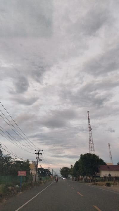 The image presents an outdoor scene along a paved road under a heavily overcast sky. The sky is dominated by a vast expanse of grey clouds, suggesting a dull day or late afternoon/early evening.

A two-lane asphalt road extends into the distance, marked by a solid white line on the left edge and dashed yellow lines in the center. On the left side of the road, multiple utility poles are visible, supporting a dense network of power lines that traverse the frame. Below these lines, lush green trees and bushes form a dense border. A blue sign with a red rectangular insert is visible on the left; the white text within the red rectangle appears to read "DOY CLUB" with an arrow pointing to the right. A yellow diamond-shaped sign is also present further along the left shoulder.

On the right side of the road, more trees line the path, behind which a building with a reddish-brown roof can be seen. Two tall, red and white lattice communication towers stand against the cloudy backdrop, one prominent in the mid-ground and another smaller one further back.

A motorcycle with a rider and a passenger is visible on the road, heading away from the viewer. Distant headlights suggest the presence of other vehicles. The overall dim lighting, influenced by the dense cloud cover, indicates a time of day transitioning towards evening. The scene captures a common roadside environment in Sukadamai, Indonesia, featuring typical utility infrastructure and vegetation.
