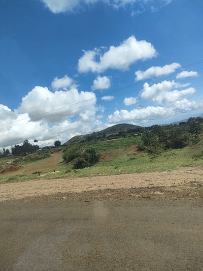 The image depicts a landscape in Kisima Ward, Kenya. In the foreground is a paved road. Beyond the road, the terrain transitions to a mix of dirt and grass. A hillside rises in the background, covered in vegetation, including what appears to be crops. The sky is blue with scattered white clouds. Power lines are visible across the upper portion of the image.
