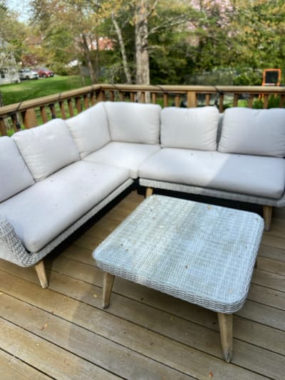 A white wicker sectional sofa and a matching coffee table are situated on a wooden deck. The sofa consists of two pieces, forming an L-shape. The coffee table is positioned in the center of the L. The deck is made of horizontal wooden planks. The scene appears to be in a backyard, as leafy trees can be seen in the background. The location is Rye, New York, which is a town on Long Island Sound, known for its beaches and historic charm. The coordinates (40.952, -73.708) place the scene in a residential area of the town. There are no people present in the image.