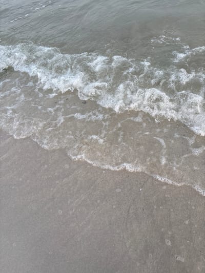 The image shows a close-up, high-angle view of a shoreline in Ajman, United Arab Emirates. Gentle waves are breaking on a sandy beach. The water is a muted gray color, and the sand is light brown with a slightly wet appearance near the water's edge. Small bubbles and foam are visible on the surface of the receding water and on the sand. The scene is devoid of any people or man-made objects, focusing solely on the natural interaction between the sea and the land. The lighting suggests an overcast or early morning/late evening time.