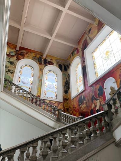 A view up a stairwell reveals a complex architectural space. A mural covers the walls, depicting historical figures and scenes. Skylights punctuate the painted surfaces, providing natural light. Below, stained glass windows add color and detail. A geometrically patterned stairway climbs, its design contributing to the overall visual interest. The ceiling is grid pattern.