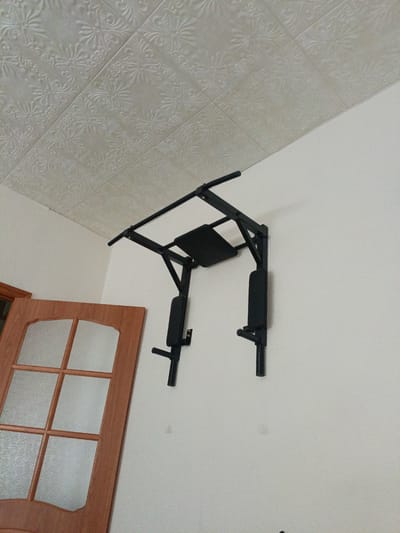 A black multi-functional pull-up bar is mounted on a white wall. The pull-up bar features a horizontal bar at the top, angled support brackets, and padded armrests and back support. To the left of the pull-up bar, a wooden door with six frosted glass panes is visible. The ceiling above is covered with decorative textured tiles. The scene is indoors, in what appears to be a room.