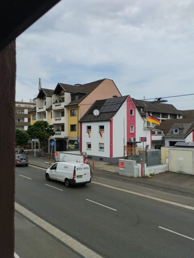 The image depicts a street scene in Troisdorf, Germany, under an overcast sky. In the foreground, a grey sedan is parked on the right side of the road, facing away from the viewer. Further down the road, a white Mercedes-Benz Sprinter van with the branding "AKOZA" is in motion, heading towards the background. The van has a license plate that appears to be "K-AZ 1037." On the right side of the street, several buildings are visible. One building is painted white with red vertical stripes and features a flag of Germany on a flagpole. Another building, adjacent to it, has a sign that reads "Bedachungen und Wa....." which likely indicates a roofing business. The buildings appear to be commercial and residential structures, with some showing solar panels on their roofs. To the left, a row of apartment buildings with balconies and yellow accents is present. Trees line the sidewalk between the road and the buildings. A blue traffic sign with a white arrow and a circle is also visible. The overall environment suggests a typical European urban or suburban setting.