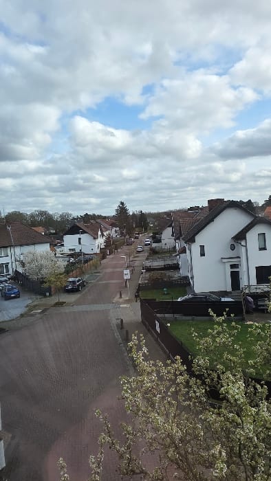 The image presents an elevated, oblique view of a residential street in Heusden, Belgium, under a partly cloudy sky with prominent cumulus formations and visible patches of blue. The street, paved with reddish-brown interlocking pavers, extends into the mid-distance where it appears to curve right. On both sides, two-story detached houses with predominantly white facades and dark, gabled roofs are visible.

In the lower left foreground, two blue vehicles, a hatchback and an SUV, are parked on a paved area adjacent to the street. Further along the left side of the street, a dark sedan is parked, and a white blossoming tree stands. On the right side, a manicured lawn is visible behind a dark brown privacy fence, with a prominent tree exhibiting white blossoms partially obscuring the lower right foreground. Beyond this fence, a two-story white house with a dark roof and a circular window detail is prominent.

Further down the street, additional parked vehicles, including several white vans or cars, are observed. Streetlights and a street sign are discernible along the roadway. The background indicates further residential development transitioning into a treelined horizon. No individuals are visible.