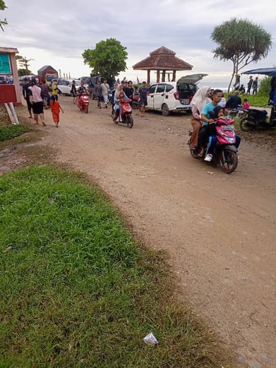 The image shows a dirt road in Pameungpeuk, Indonesia. People are walking, riding motorcycles, and driving cars. Several motorcycles are parked on the side of the road. A woman and a young girl are riding a red motorcycle on the right side of the road, going in the same direction as several other motorcycles. There is a white car parked on the right side of the road, and a group of people are gathered at the side of the road in the background. There is a small red building on the left side of the road. There is a lot of green grass on the left side of the road. The road is dusty and dry. The sky is cloudy. The coordinates for this location are -7.668, 107.694.