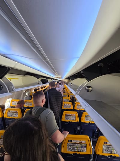 The image captures the interior of a passenger airplane cabin, likely a Ryanair flight given the yellow seatbacks, as passengers queue in the aisle. The overhead compartments are open, with some luggage visible inside. The lighting suggests the aircraft may be on the ground, possibly at John Paul II International Airport Kraków–Balice (KRK) in Balice, Poland, as indicated by the presence of the text "EXIT WYJSCIE" on a sign. The aisle is crowded with people, some standing and others seated, waiting to disembark or board. The perspective is from a passenger in the aisle, looking forward towards the front of the plane.