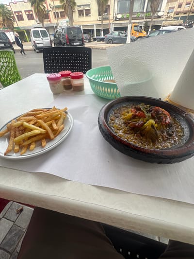 The image captures an outdoor dining scene, rotated vertically, viewed from a seated perspective at a table overlooking a street. In the foreground, a traditional earthenware tagine, filled with a rich, saucy stew of vegetables (including tomatoes and possibly other elements), takes center stage. Alongside it, a white oval plate holds a serving of golden French fries. Three small spice shakers with red and pink lids, a stack of green plastic bowls/baskets, and a roll of paper towels are also arranged on the white paper-covered table.

The background reveals a lively street in what appears to be a town or city. Multiple vehicles are visible, including a white delivery van with "CENTRA P M A D" printed on its side, a black car, a grey car, and a distinctive small yellow vehicle. A person walks on the sidewalk. The street is lined with multi-story buildings, some featuring storefronts with glass windows. Partial text "Restaurant Glacier l'Hir..." and "Cafe" can be discerned on building signs. Several palm trees dot the streetscape.

The scene is illuminated by natural light, indicating daytime with clear weather. The presence of the tagine, palm trees, and architectural style, combined with the location context of Boufakrane, strongly suggests a Moroccan setting. A person's leg and arm are partially visible, indicating they are seated at the table.