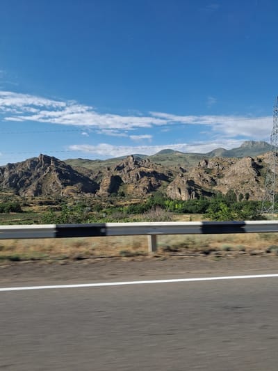 The image depicts a mountainous landscape under a clear blue sky with scattered white clouds. The foreground features a road with a white line marking the lane. A guardrail with black and white stripes separates the road from the natural landscape. In the background, rugged, rocky mountains rise, covered in dry grass and sparse vegetation. A cluster of green trees is visible in a valley between the mountains. A tall, metal electrical pylon stands on the right side of the image. The scene appears to be in or near Malishka, Armenia, suggested by the rocky terrain and vegetation typical of the region. There are no people or animals visible in the image, nor any overt actions or interactions taking place.
