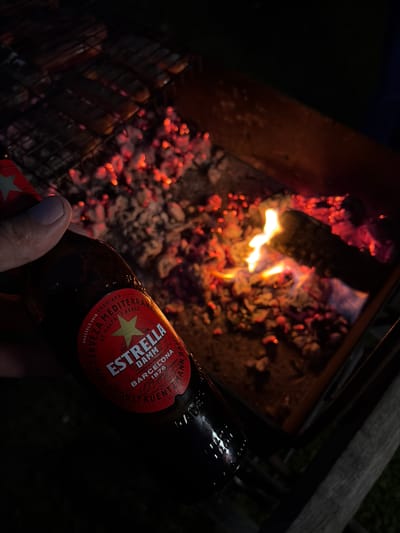 In this media file, the subject is a person holding a bottle of Estrella Damm beer in front of a glowing barbecue grill. The grill is filled with burning embers and flames, suggesting that food is being cooked or has recently been cooked. The scene is set outdoors at night, as indicated by the dark background and the artificial light from the fire and the bottle label. The person's hand is visible, holding the bottle with their thumb resting on the label, implying a relaxed and enjoyable moment.
The Estrella Damm beer bottle features a red label with gold lettering. Visible text on the label includes "ESTRELLA DAMM BARCELONA 1876", "CERVEZA MEDITERRANEA", and "BRUSSELS 2018 PREMIADA". The presence of these awards and the brand name suggests a focus on the quality and origin of the beer. The overall atmosphere conveyed is one of leisure and outdoor dining, possibly a barbecue gathering on a summer evening. The location context, "Манихино Снт, Russia," places this scene in a specific Russian dacha or cottage area. The time of day is clearly night, with the fire providing the main illumination in the immediate vicinity.