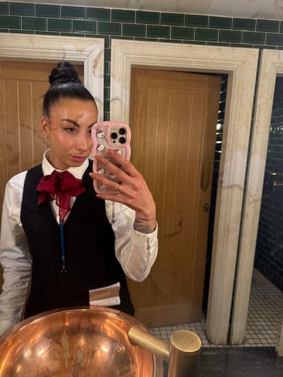 A woman is taking a selfie in what appears to be a restroom. She has dark hair pulled back in a bun, and she's wearing a white collared shirt with a black vest and a red bow tie. She's holding up a pink Hello Kitty iPhone to take the photo. Behind her is a wood-paneled door with a marble-patterned frame. To the left is green tiled wall and to the right is a copper sink. I have no information to confirm if this image was taken in Warrington, United Kingdom.