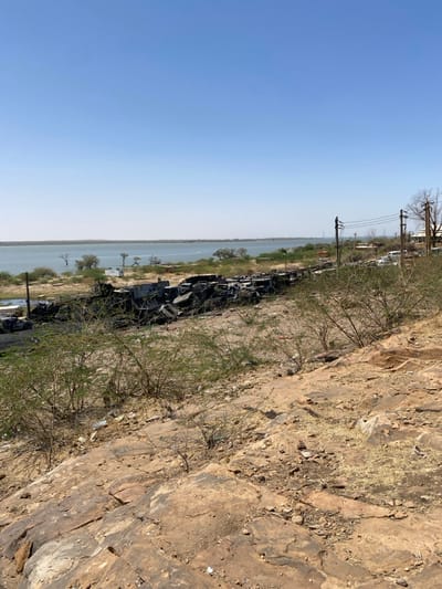 The image shows a view from a rocky hill overlooking a body of water, possibly the Nile River. The location is Tayba Alhasanab, Sudan, with coordinates 15.362, 32.525. The scene is characterized by a vast expanse of water with a clear, blue sky. The ground is covered in rocks and sparse vegetation, with a line of burnt and damaged vehicles clustered near the water's edge. Power lines stretch across the horizon, and several trees can be seen on the distant shore. The image captures a post-conflict or disaster setting.