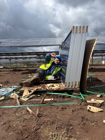 The image shows a man standing among solar panels, apparently during installation or maintenance. The setting seems to be a solar farm, potentially located in Galatro, Italy, as specified in the prompt.
**People:** A man is present, wearing a blue hard hat, a yellow reflective vest, and boots. He is holding an unidentified object in his hand.
**Subjects:** A large array of solar panels dominate the background, suggesting an industrial or agricultural installation.
**Objects:** Other than the solar panels and the object the man is holding, there is packaging material and green straps on the ground, possibly used to secure the panels during transport.
**Actions:** The man is looking at the camera, and holding an object in his hand.
**Interactions:** The man appears to be at work, interacting with the solar panel equipment and the surrounding environment.
**Environment:** The ground is brown soil, and the sky is overcast. The overall scene suggests a utility-scale solar power installation in a rural or agricultural area.