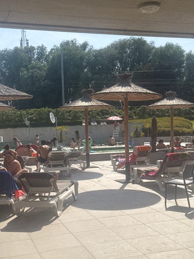 This is a photograph taken outdoors, likely during the daytime given the strong shadows cast by the objects and the bright sunlight. The scene appears to be a resort or a public pool area, with a tiled ground and several empty lounge chairs set up for relaxation. In the background, there are lush green trees and some foliage, suggesting a warm and possibly summery weather.
The central focus of the image is a pool area where several people are visible. Some are lounging on chairs, while others are in or near the water. The people are dressed in swimwear, indicating they are engaging in recreational activities related to the pool. The atmosphere seems relaxed and leisurely, with individuals enjoying the warm weather and the amenities of the location.
Notable details include several thatched umbrellas providing shade, typical of outdoor leisure spaces. The light suggests it's either morning or late afternoon. The overall impression is one of a pleasant day at a vacation spot. There is no visible text in the image.