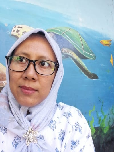 The image is a selfie of a woman wearing a light blue hijab and a white shirt with a blue floral pattern. She is also wearing a silver flower-shaped brooch on her hijab. She has dark eyes and is wearing black-rimmed glasses. The background is a mural of an underwater scene, with a sea turtle and fish painted on a blue wall. The woman is standing in front of the mural, which appears to be located outdoors. The overall scene suggests a casual and artistic environment.
