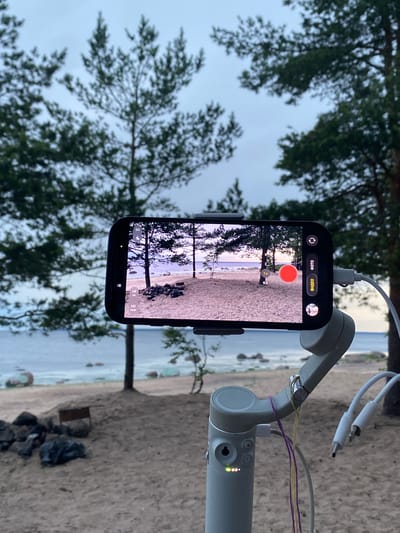 An iPhone is mounted on a gimbal and is filming a beach scene at dusk in Primorsk, Russia. The iPhone displays a view of a sandy beach with scattered rocks and trees. In the foreground, the gimbal is visible with charging cables attached and colorful strings tied to it. The sea is visible in the background, with a cloudy, overcast sky. There are no people in the image.