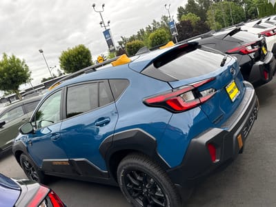 In Puyallup, United States of America, a row of Subaru Crosstrek vehicles are parked on an asphalt lot. The primary vehicle in the foreground is a blue Crosstrek, viewed from the rear passenger side. It features black plastic cladding around the wheel wells and lower body panels, a roof rack with yellow accessories, and a "CROSSTREK" badge on the lower door. The rear of the vehicle displays distinctive red taillights and a prominent black bumper with integrated amber reflectors. Behind the blue Crosstrek, other Crosstreks are visible in black and white, lined up in the lot. In the background, Subaru banners hang from light poles, and a line of trees forms the backdrop against an overcast sky. The scene appears to be at a car dealership.