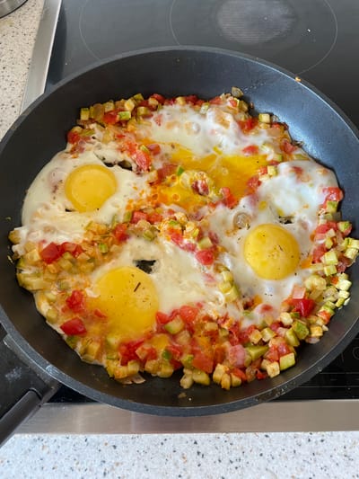 A top-down shot shows a frying pan containing eggs cooked with diced vegetables. The pan is black and sits on a black stovetop with a light speckled countertop surrounding it. The eggs are mostly cooked, with some yolks intact and some broken. The diced vegetables appear to be primarily zucchini and tomato. The scene appears to be indoors. The location is Бордуки, Russia.