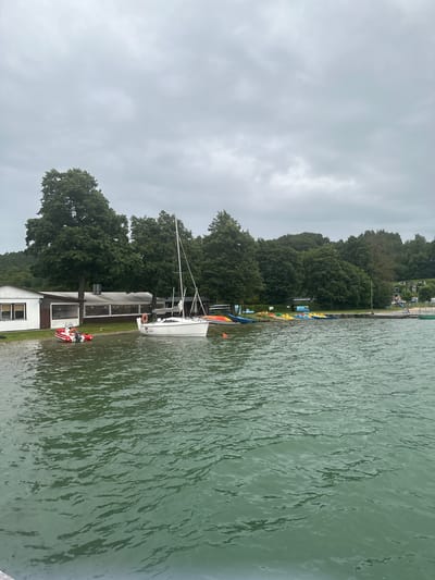 The image depicts a waterfront scene in Chmielno, Poland, under an overcast sky. In the foreground, choppy green water fills the frame, with visible ripples and waves. Along the shore, a white building with a dark roof and large windows is partially submerged. Adjacent to it, a restaurant or event hall with a covered outdoor seating area is visible.
Several vessels are present: a white sailboat with a black sail is moored near the shore. To its left, a small red inflatable boat with an outboard motor is partially in the water. Further down the shoreline, a collection of colorful paddle boats and kayaks are lined up. A wooden pier extends into the water in the distance.
The background is dominated by a dense line of lush green trees. Beyond the trees, several small houses or cabins are visible, suggesting a residential or resort area. The overall impression is of a calm, slightly gloomy day at a lakeside recreational spot. There are no people clearly visible interacting in the scene.