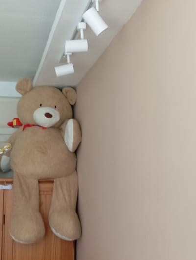 The image shows a large, tan teddy bear lying down. The bear has a red scarf around its neck. It is positioned on top of a wooden dresser or cabinet. In the background, there are white track lights mounted on a ceiling or wall. The scene appears to be indoors, possibly a bedroom or living space. The lighting suggests it might be daytime, but no external cues are visible. The overall mood is relaxed and perhaps a bit whimsical due to the presence of the oversized stuffed animal. There is no visible text in the image.