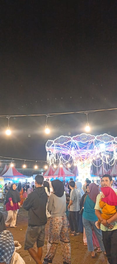 The image depicts a bustling night scene at what appears to be a local fair or amusement park in Banding Agung, Indonesia. A prominent feature is a brightly lit carousel or swing ride adorned with numerous colorful lights and streamers. Several strings of lights are suspended overhead, illuminating the gathering.
In the foreground, a crowd of people is visible, suggesting a lively atmosphere. To the left, a woman wearing a dark hijab and a magenta top stands with her arms crossed, looking to the side. Beside her, a man in a dark hoodie and grey shorts is seen from the back. Further right, a person in a grey top and patterned pants also faces away from the camera. On the far right, a man in a light green shirt holds a child wearing a red hijab and an orange outfit.
In the mid-ground, more people are gathered, and red and white striped tents are visible, likely stalls or attractions at the fair. The ground appears to be dirt or sand. The overall impression is one of a community event taking place at night, characterized by light, activity, and a sense of local festivity.