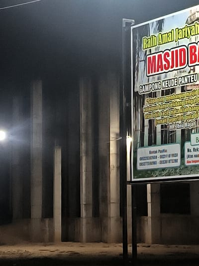 A nighttime, close-up shot reveals a partially constructed building in Keudepanteubreuh, Indonesia. Concrete pillars rise from the ground. A large, worn sign advertising the "Masjid B" is situated in front of the construction. The sign contains text in an Indonesian dialect, including phone numbers and religious text. The surrounding environment is dark, with minimal ambient light.
