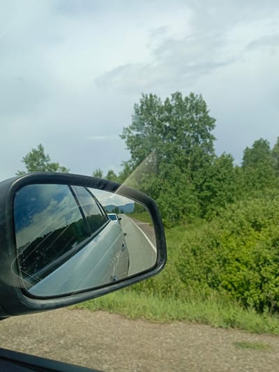 The image, set in Usakly, Russia, is captured from the interior perspective of a vehicle, focused on the exterior passenger-side mirror. The sky is overcast with a light gray hue. The foreground displays an asphalt road with a white demarcation line. Adjacent to the road, dense green vegetation, comprising bushes and deciduous trees, extends into the midground and background. Distant mountains are discernible through the vehicle's window and in the mirror's reflection. The black-framed mirror exhibits surface abrasions and streaks, reflecting the cloudy sky, distant landscape, a portion of the originating light-colored vehicle, and a white vehicle positioned ahead on the road. No individuals are depicted.