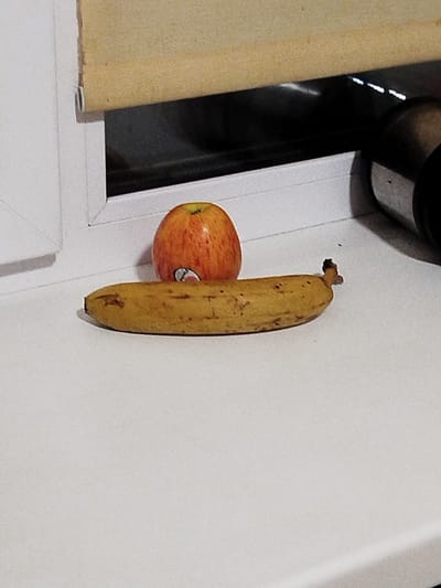 On a white windowsill, a browning banana lies horizontally in the foreground. To the left and slightly behind the banana, an apple with red and yellow skin is positioned upright. Behind the fruits, a dark reflective surface is visible, possibly a window. Above the window, a beige roller blind hangs down. To the right of the fruits and in the background, a portion of a dark cylindrical object, likely a thermos or bottle, is visible. The scene is set indoors.