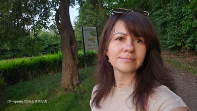 A woman with brown hair, wearing a light-colored t-shirt and glasses perched on her head, takes a selfie outdoors. She is standing in a park with a tree to her left and a path to her right. In the background, there is a signpost with some text and graphics, surrounded by lush green trees and bushes. The scene suggests a pleasant day in a natural setting, likely Bannewitz, Germany, given the timestamp on the image, which reads "19 червня 2026 р. 9:03 PM". The lighting indicates it is late afternoon or early evening.