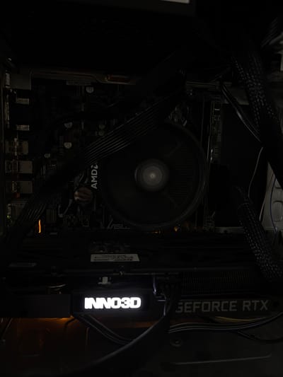 This is a close-up, dimly lit photograph taken inside a computer case, showing internal components. The focal point is a graphics card with the prominent branding "INNO3D" illuminated in white text, and further to the right, "GEFORCE RTX" is also visible. The computer's motherboard is partially visible, with a large CPU cooler fan dominating the center. Black cables are routed throughout the case, obscuring some of the components. The overall scene is dark, with some internal LEDs providing subtle illumination, including a faint orange glow on the left side of the motherboard and the white illumination from the graphics card branding. The location of Mülheim an der Ruhr, Germany is not depicted in this image.