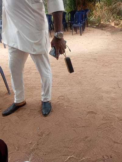 The image displays an outdoor scene in Okpatu-Ukehe, Nigeria, on reddish-brown earthen ground marked with numerous footprints. A male subject is partially visible, wearing a white long-sleeved tunic over white trousers and dark blue or black slip-on loafers adorned with an "H" emblem on the vamp. A prominent silver-toned wristwatch is on his right wrist, and a ring is on his right ring finger. His right hand holds a black smartphone, with a small, rectangular black electronic device, likely a power bank or portable speaker, dangling from it by a cable. In the background, a stack of approximately seven blue plastic chairs is visible, behind which sparse, dry vegetation, including tall grasses, is present.