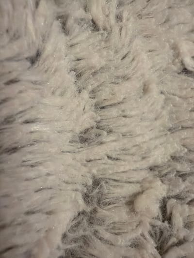 The image presents a macro-level view of a textured, fibrous material. The primary subject is a dense aggregation of tangled and matted fibers, predominantly off-white and light gray in color. Darker grey and black tones are visible in the interstices, suggesting depth or an underlying surface. Individual fibers vary in length and orientation, creating an irregular, plush texture. Numerous small, bright, specular points of light, resembling glints or sparkles, are scattered across the surface of the fibers. No people, specific objects beyond the textile, or actions are discernible within the frame. The immediate environment is solely defined by the fibrous material under close examination.