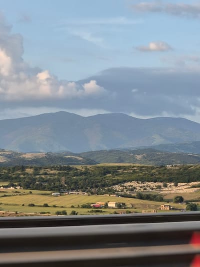 The image captures a vast rural and mountainous landscape, attributed to the city of Moshtanets, Bulgaria, viewed from behind a dark grey, two-tiered guardrail in the immediate foreground, suggesting an elevated vantage point such as a road or bridge.

The scene is dominated by layers of landforms under a partially cloudy sky. In the distant background, several large, hazy, dark green-blue mountains with undulating peaks span the horizon. Closer, but still extending towards the midground, are rolling hills covered in varying shades of green vegetation and some patches of lighter, drier grass or earth.

The midground features cultivated fields in shades of yellow-green, interspersed with numerous clusters of dense green trees. Several small, scattered buildings or houses with light-colored walls and dark roofs are visible within these fields and among the trees. A prominent feature on a distant hillside is a large area densely covered with hundreds, possibly thousands, of small, regularly spaced white or light-colored rectangular markers, arranged in neat rows along the contours of the slope, indicative of a large cemetery or memorial site.

The sky occupies the upper portion of the frame, displaying a pale blue hue with scattered cumulus clouds. Some clouds are bright white from sunlight, while others are darker grey, indicating varying light conditions. The overall lighting suggests either late afternoon or early morning, with soft illumination across the landscape. No human figures or active interactions are discernible.