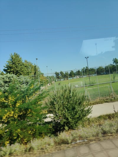 The image shows an outdoor scene with a clear blue sky. In the foreground, blurred green foliage with yellow flowers is visible, suggesting movement. Beyond the foliage, a fenced sports field, likely a soccer field, is present with goalposts. Several tall light poles stand on the field, indicating it may be used for evening games. In the background, a line of trees and some buildings are visible. The reflection on a window or glass surface reveals text that appears to be in French, possibly indicating "ISSUE DE SECOURS" which means "emergency exit". The overall environment suggests a park or sports complex in an urban or semi-urban setting, with the mention of Larissa, Greece, placing it geographically. A person on a yellow vehicle is visible on the field.